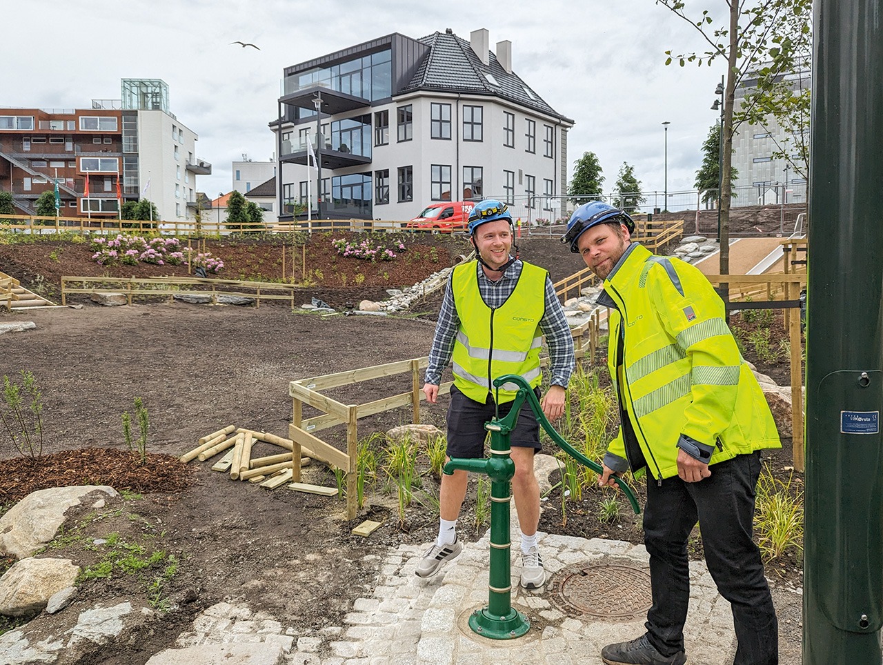 Under den manuelle vannpumpen er det en kum som kan samle opp ca 1000 liter vann. Fra venstre prosjekt ingeniør Lars Ottestad og driftsleder Anton Komarov fra Consto AS. De midlertidige tre-gjerdene skal tas ned når plantene har satt seg.